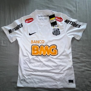 Santos Neymar Jr 11 Nike fotbollströja - Vit Santos fotbollströja från Nike med Neymar Jr och nummer 11 på ryggen. Tröjan har korta ärmar, Dri-Fit-material och flera tryckta sponsorer. Snygg klubbsköld på bröstet och klassisk passform. Perfekt för dig som älskar brasiliansk fotboll och Neymar.
