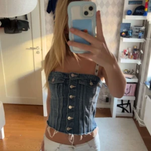 Jeans korsett-topp med knappar - Säljer en snygg korsett-topp i jeans med knappar framtill och råa kanter nertill. Toppen är croppad och har en cool denimlook med fickdetaljer upptill. Perfekt för dig som vill sticka ut med en trendig och edgy stil.