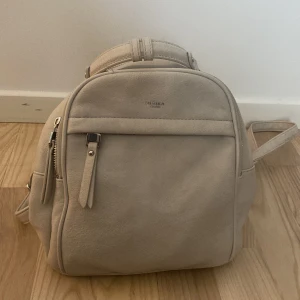 Beige ryggsäck från Ulrika Design - Snygg beige ryggsäck från Ulrika Design med stilren form och flera praktiska fack. Väskan har justerbara axelremmar, dragkedjor i silver och är tillverkad i mjukt skinnliknande material. Perfekt för dig som vill ha en clean och enkel look.