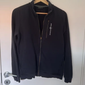 Svart jacka från Sail Racing - Svart jacka från Sail Racing med hel dragkedja framtill och en bröstficka med logga. Jackan har en stilren look, hög krage och är tillverkad i ett mjukt material som känns skönt mot huden. Perfekt för dig som gillar sportig och clean stil.