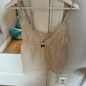 Beige linnetopp från Odd Molly - Supersöt beige linnetopp från Odd Molly i storlek S, med tunna axelband, spetsdetaljer längs kanterna och en liten vinröd rosett framtill. Broderad logga på ryggen och justerbara axelband. Kommer tyvärr inte till användning. 