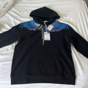 Marcelo Burlon svart hoodie med blå vingar - Säljer en svart hoodie från Marcelo Burlon med sjukt snyggt blått och silverfärgat vingmönster över axlar och rygg. Tröjan har huva med snörning och stor magficka framtill. Perfekt för dig som vill sticka ut med en unik design.