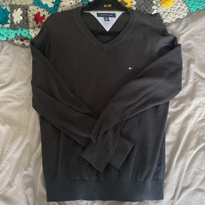 Svart v-ringad tröja Tommy Hilfiger - Svart v-ringad tröja från Tommy Hilfiger med diskret logga broderad på bröstet. Tröjan har långa ärmar, ribbade muddar och är gjord i mjuk bomull. Perfekt för dig som gillar stilrena och klassiska plagg med en modern touch.