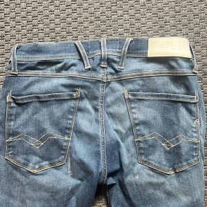 Blå Replay jeans med slitningar - Säljer ett par blå jeans från Replay med snygga slitningar och ljusare partier framtill. Klassisk femficksmodell med Replay-logga på fickan och bak på linningen. Jeansen har raka ben och kontrastsömmar som ger en schysst look. Använd fåtal ggr