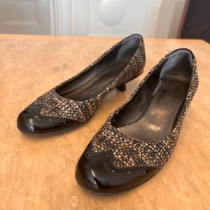 Mönstrade pumps med taxklack, stl 37 - Pumps från Tamaris med låg klack i storlek 37. Skorna har ett vävt, flerfärgat mönster i svart, beige och brunt samt svarta lackdetaljer vid tå och häl. Rund tå och klassisk siluett som ger en chic touch till din outfit. Har endast använts inomhus.