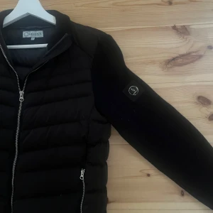 Svart pufferjacka från Lagency - Svart pufferjacka från Lagency med quiltad front och ärmar i mjuk ullmix. Jackan har två dragkedjefickor, hel dragkedja framtill och en stilren patch på ena ärmen. Perfekt för dig som gillar en clean och modern look.