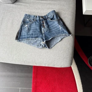 Blå jeansshorts med knappar - Snygga blå jeansshorts med hög midja och råa kanter. Framtill finns fyra synliga knappar och slitna detaljer för en cool look. Svarta paljettpaneler på sidorna ger extra edge. Shortsen har klassiska fickor både fram och bak.