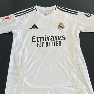 Real Madrid Mbappé matchtröja 24/25 - Snygg vit Real Madrid fotbollströja från Adidas med Mbappé och nummer 9 på ryggen. Tröjan har svarta detaljer, klubbmärke på bröstet, La Liga-märke på ärmen och klassiska Adidas-ränder på axlarna. Tillverkad i lätt och ventilerande material.