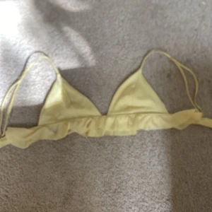 Gul bikiniöverdel med volangdetalj - Söt gul bikiniöverdel med trekantskupor och volang längs underkanten. Justerbara axelband och metallspänne i ryggen för perfekt passform. Perfekt för strandhäng och poolparty och badhus den har små gråa fläckar som går bort i tvätten