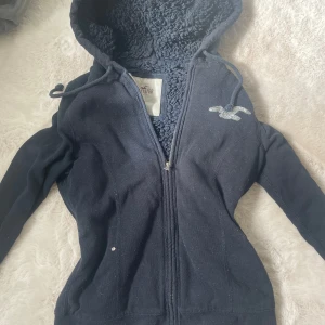 Vintage Hollister zip up - Mysig mörkblå teddyjacka från Hollister med dragkedja och huva. Insidan är fodrad med fluffigt teddyfoder och jackan har snörning i huvan samt Hollister-logga på bröstet. Perfekt för kyliga dagar och riktigt skön att ha på sig.