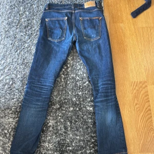 Nudie Jeans blå slim fit jeans - Snygga blå jeans från Nudie Jeans med klassisk femficksdesign och kontrastsömmar. Jeansen har en slim passform och raka ben, med tydliga slitningar och Nudie-logga på bakfickan. Perfekta för dig som gillar stilrena och tidlösa jeans.