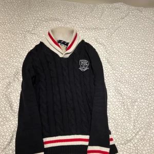 Morris kabelstickad tröja med krage - Snygg kabelstickad tröja från Morris i marinblått med vit och röd ribbad krage samt muddar. Tröjan har lång ärm, broderad logga på bröstet och Union Jack-detalj på ärmen. Perfekt för dig som gillar preppy stil och vill sticka ut lite extra.