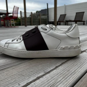 Valentino Garavani sneakers vit/vinröd - Snygga Valentino Garavani sneakers i vitt skinn med bred vinröd detalj över mitten och coola nitar på hälen. Klassisk rund tå, platt sula och grå snörning. Perfekta för dig som vill ha en lyxig och stilren look med edge. säljes utan box