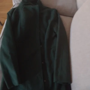 Mörkgrön kappa från Atmosphere - Snygg mörkgrön kappa från Atmosphere i klassisk rak modell med knappar framtill och hög krage. Perfekt för dig som vill ha en stilren och enkel jacka med lång ärm och tidlös look. Passar till många olika outfits och håller dig varm under kyliga dagar.