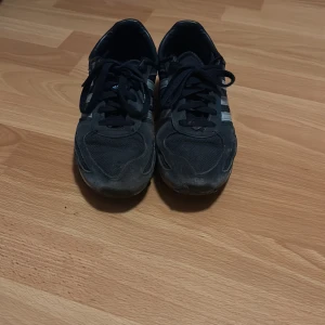 Svarta Adidas sneakers med vita detaljer - Svarta Adidas sneakers med klassiska tre vita ränder på sidorna och vit häl med Adidas-logga. Skorna har snörning, rund tå och platt sula. Ovandelen är i en mix av syntet och mocka för en sportig look. Perfekta för dig som gillar streetstyle. Djurgården 