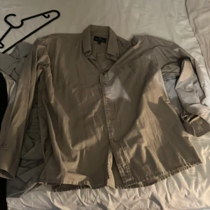 Beige skjorta från Rappson i bomull - Säljer en klassisk beige skjorta från Rappson i storlek L. Skjortan är tillverkad i bomull, har lång ärm, knappar framtill och en stilren krage. Perfekt för dig som gillar en enkel och clean look.