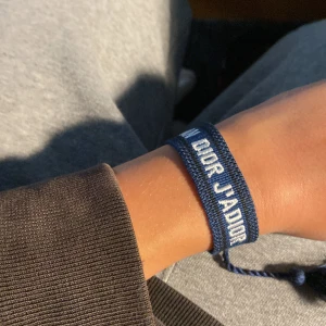 Dior armband - Snyggt blått armband från Christian Dior med texten 'J'Adior' broderad i vitt. Armbandet är gjort i ett mjukt tygmaterial och har justerbar knytning för perfekt passform. Perfekt accessoar för att lyfta din outfit med lite lyxig vibe.