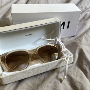 Chimi solglasögon Ecru med guld - Snygga solglasögon från Chimi i modellen Ecru. Bågarna är transparenta med en beige ton och har guldiga detaljer på insidan av skalmarna. Glasen är bruna och har en lätt fyrkantig form. Kommer med originaletui och box.