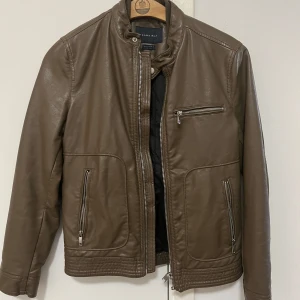 Brun skinnjacka från Zara Man - Snygg brun skinnjacka från Zara Man med klassisk rak passform. Jackan har ståkrage med tryckknapp, tre dragkedjefickor framtill och detaljerade sömmar vid nederkant och ärmslut. Perfekt för dig som gillar stilren och tidlös look.