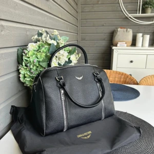 Svart handväska Zadig & Voltaire - Snygg svart handväska från Zadig & Voltaire i strukturerat skinn med silverfärgade dragkedjor och detaljer. Väskan har två rejäla handtag, innerfack med dragkedja och ikonisk logga framtill. Perfekt storlek för allt du behöver ha med dig.