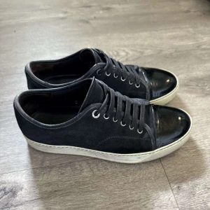 Svarta sneakers från Lanvin med lackad tå - Snygga svarta sneakers från Lanvin med ovandel i mocka och en glansig, lackad tå. Skorna har vita sulor och klassisk snörning med metallöljetter. Perfekta för dig som gillar en stilren men ändå unik look. Skriv vid funderingar?