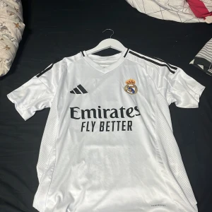 Real Madrid vit matchtröja Adidas S - Säljer en vit Real Madrid fotbollströja från Adidas i slim fit, storlek S. Tröjan har svarta detaljer och klubbmärke på bröstet, samt 'Emirates Fly Better' tryckt framtill. Tillverkad i lätt och ventilerande polyester med korta ärmar och klassisk v-ringad krage.