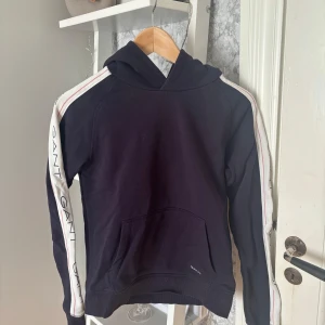 Marinblå  hoodie från GANT med logoband - Marinblå  hoodie från GANT med vita logoband och röda detaljer längs ärmarna. Klassisk huva och magficka framtill. GANT-logga broderad på bröstet och längs ärmarna. Skön passform och tillverkad i mjuk bomull, perfekt för chill dagar.