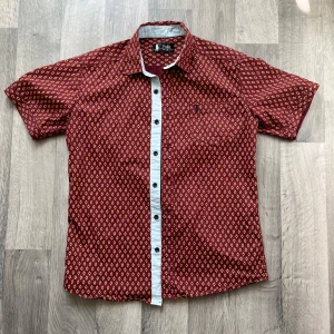 Mönstrad kortärmad skjorta Polo Ralph Lauren - Snygg kortärmad skjorta från Polo Ralph Lauren i vinrött med gult geometriskt mönster. Skjortan har vit knappslå med svarta knappar, klassisk krage och bröstficka med broderad logga. Perfekt för dig som gillar färg och mönster.