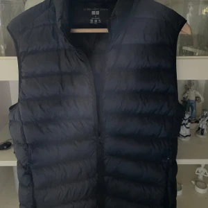 Uniqlo väst - Säljer en svart, superlätt dunväst från Uniqlo Ultra Light Down. Västen är quiltad med horisontella sömmar, har dragkedja framtill och hög krage. Perfekt att slänga över en hoodie eller tröja när det är kyligt ute. Kommer med tillhörande förvaringspåse. I nytt skick.