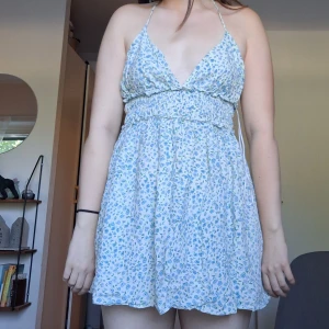 Blommig kort sommarklänning med v-ringning - Söt kort klänning med ljusblått blommönster på vit botten. Klänningen har v-ringad hals, smala axelband och omlottkänsla upptill. Smockad midja ger en skön passform och den är ärmlös. Perfekt för varma dagar och har en flowy, lätt känsla.