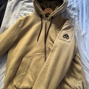 Beige hoodiejacka från Moose Knuckles - Snygg beige hoodiejacka från Moose Knuckles med dragkedja, huva och svarta snören. Jackan har quiltat foder och en ikonisk logga på ärmen. Perfekt för dig som vill ha en stilren och bekväm jacka med streetkänsla.knappt använd,såklart ÄKTA.pris kan diskuteras 