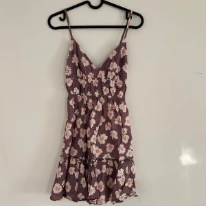 Lila blommig kortklänning med volang - Supersöt lila kortklänning med blommor, smala axelband och v-ringad hals. Klänningen har smock i midjan och volang nedtill. Materialet känns lätt och luftigt. Storlek xs, liten i storleken.