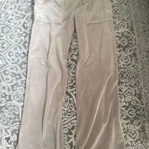 Beige velourbyxor Juicy Couture - Mysiga beige velourbyxor från Juicy Couture med breda ben och stora fickor både fram och bak. Byxorna har broderad JC-logga på ena fickan och elastisk midja för extra komfort. Perfekta för en chill och trendig look.