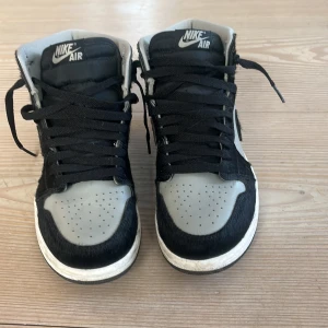 Nike Air Jordan 1 High svart/grå - Nike Air Jordan 1 High sneakers i svart och grått med pälsdetaljer och klassisk snörning. Skorna har rund tå, vit platt sula och silverfärgad Air Jordan-logga på sidan. Materialmix av skinn och päls ger en unik vibe. Perfekt för dig som vill sticka ut med din sneakerstil.