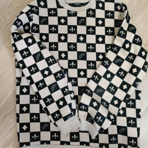 Svartvit rutig sweatshirt Icono Couture - Säljer en svartvit sweatshirt från Icono Couture med rutigt mönster och coola detaljer som logga och liljor på varje ruta. Tröjan har rund hals och långa ärmar, perfekt för dig som gillar statement-plagg och streetstyle.