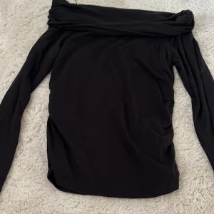 Svart offshoulder topp Gina Tricot - Svart offshoulder topp från Gina Tricot med långa ärmar och snygg rynkdetalj på sidorna. Toppens material är mjukt och stretchigt, perfekt för en trendig och stilren look. Passformen är tight och axelband saknas för en clean offshoulder-stil. Inte använt en ända gång bara att lappen är borta.