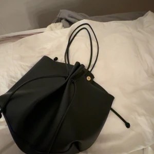 Svart bucket bag i skinnimitation - Snygg svart bucket bag med dragsko och långa axelband. Väskan har minimalistisk design och är gjord i slät skinnimitation. Perfekt storlek för att få plats med allt du behöver och passar till många olika outfits.