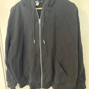 Svart hoodie med dragkedja från H&M - Svart hoodie från H&M i storlek XL med huva och snörning. Tröjan har en praktisk dragkedja framtill och två stora fickor. Materialet är mjukt och känns skönt mot huden, perfekt för chill dagar. Klassisk loose passform och långärmad design. Använd fåtal gånger.