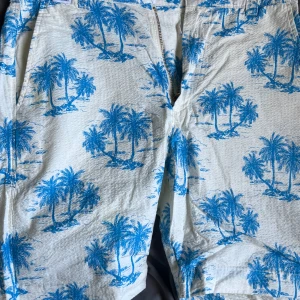 Vita shorts med blått palmmönster Rotation - Snygga vita shorts från Rotation med blått palmmönster över hela tyget. De har dragkedja fram, sidofickor och bakfickor med knappar. Perfekta för sommaren med sin lätta bomullskänsla och tropiska vibe.