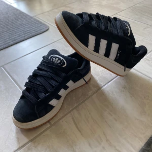 Adidas Campus 00s  - Säljer ett par Adidas Campus sneakers i svart mocka med klassiska vita ränder och logga på hälen och plösen. Skorna har snörning, platt sula i gummi och rund tå. Perfekt för dig som gillar retrostil och vill ha något som sticker ut med snygg kontrast mellan svart och vitt. Skriv för mer bilder! Dessa är endast använda under arbetstid och är i bra skick. Ända skadan är en liten skada (se bild 3)
