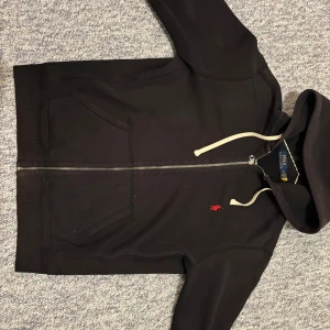 Svart hoodie från Polo Ralph Lauren - Svart hoodie från Polo Ralph Lauren med dragkedja och vit snörning i huvan. Klassisk röd Polo-logga broderad på bröstet. Tröjan har två fickor framtill och är tillverkad i mjuk bomull. Perfekt för chill dagar eller när du vill ha en clean look.
