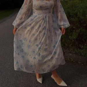 Blommig långklänning med puffärmar - Romantisk långklänning i ljus, transparent tyg med blå blommigt mönster. Klänningen har puffiga långärmar, markerad midja och fyrkantig urringning. Materialet känns som lätt polyester eller chiffong, perfekt för sommarkvällar. Söt och feminin vibe!