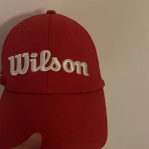 Röd Wilson keps med broderi - Snygg röd keps från Wilson med vit broderad logga framtill och WS-märke på sidan. Kepsen har perforerade paneler för extra ventilation och justerbar snapback baktill. Perfekt för dig som gillar sportig stil och vill sticka ut med en klassisk look. Har inte använt den. 