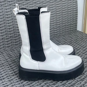 Vita chunky chelsea boots - Snygga vita chelsea boots med svart resår på sidorna och kraftig svart platåsula. Boots med rund tå och dragflik bak. Perfekta för dig som vill sticka ut med en modern och edgy look.Använda 1-2ggr, mkt bra skick. Grova boots i vitt lack på ovansidan och svart sula.  Storlek 37, normal i storlek.