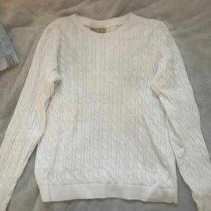 Vit kabelstickad tröja - Säljer en vit kabelstickad tröja med rund hals och långa ärmar. Tröjan har ett klassiskt flätmönster och är gjord i mjukt material som passar perfekt för kyliga dagar. Enkel och stilren design som funkar till jeans eller kjol.