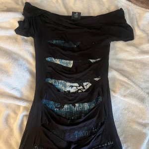 Svart kortklänning Alchemy England - Cool svart kortklänning från Alchemy England med offshoulder och slitsade detaljer framtill. Tryck med vingar och text på bröstet, samt mystiska grafiska mönster. Materialet är mjukt och stretchigt, perfekt för dig som gillar edgy stil.