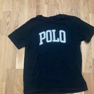Svart POLO t-shirt från Ralph Lauren - Svart t-shirt från Ralph Lauren med stor vit POLO-print på bröstet. Klassisk rund hals och korta ärmar. Tillverkad i mjuk bomull som är skön mot huden. Perfekt för dig som gillar stilrena och sportiga vibes.
