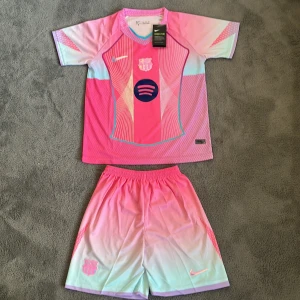 Barça new kit (REP) - Säljer ett Barcelona x Nike fotbollströja och shorts set i Dri-FIT material. Tröjan är kortärmad med coola grafiska linjer och FCB-logga på bröstet. Färgen är en mix av rosa, ljusblå och lila toner med gradient-effekt. Perfekt för dig som vill sticka ut på planen.