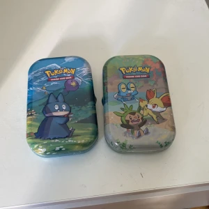 Pokémon plånbok i metall med motiv - Säljer två Pokémon plånböcker i metall med färgglada motiv av olika Pokémon-karaktärer. Perfekt för att förvara samlarkort eller småsaker. Ena har blå och gröna toner med Munchlax och Drifloon, andra har beige och grönt med Chespin, Fennekin och Froakie samt Pikachu på baksidan.