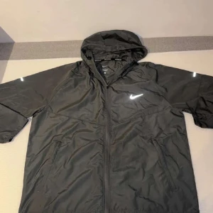 Nike Miler Vindjacka - Säljer denna gråa Miler vindjacka från Nike med huva och dragkedja hela vägen. Jackan har reflekterande detaljer på ärmar och bröst, samt två sidofickor. Perfekt för löpning eller när du vill ha en lätt jacka med sportig vibe.